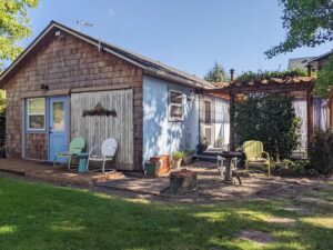 A brown shingled cottage with blue door, one blue side, and a patio with vintage colorful chairs.